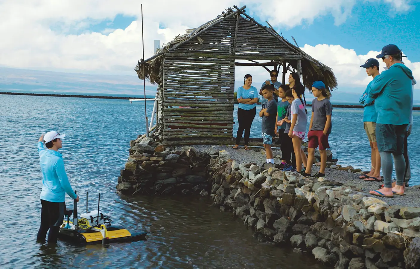At a dock on a fishpond in Molokai, Hawaii, a BYU student explains an autonomous water drone to a group of younger students.