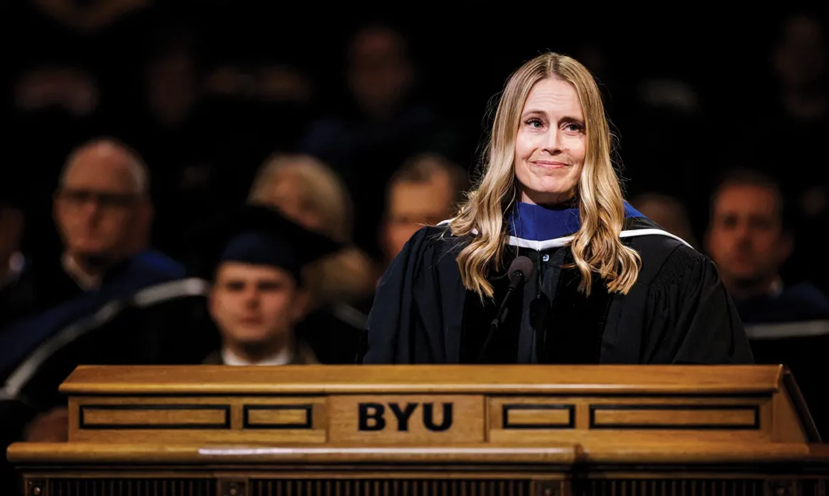 Hillary Nielsen stands at the BYU podium in commencement robes, looking off camera and smiling.