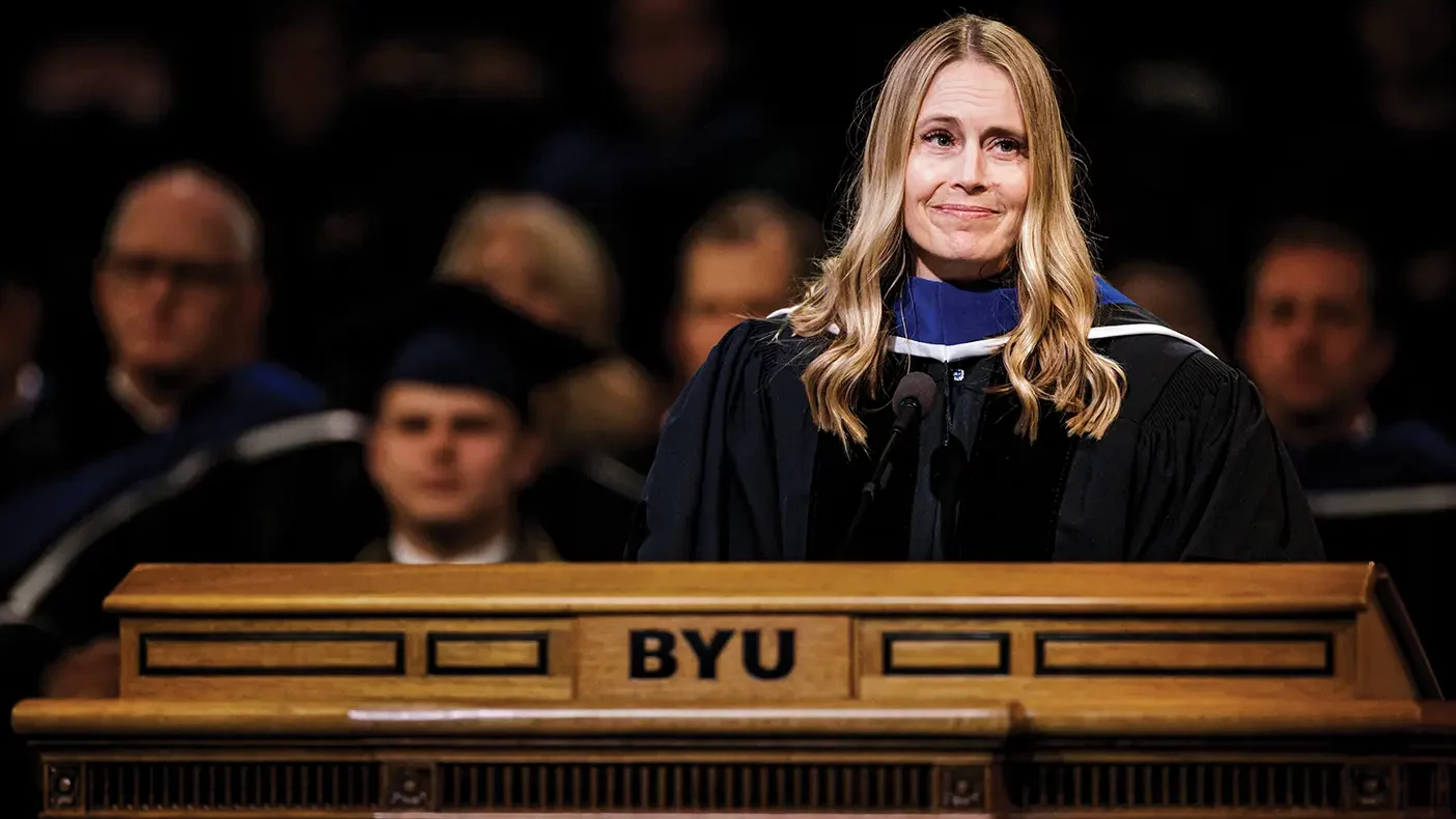 Hillary Nielsen stands at the BYU podium in commencement robes, looking off camera and smiling.