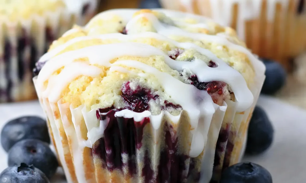 A lemon blueberry muffin drizzled in frosting sits on a white plate, garnished with seven blueberries.