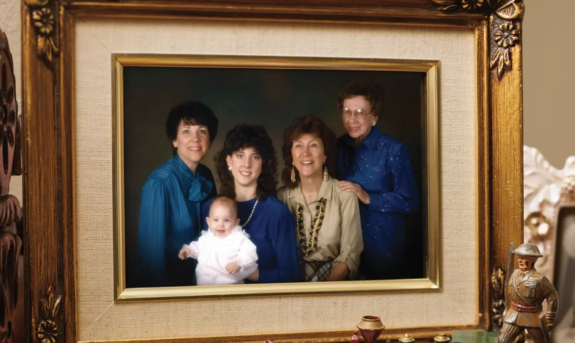 In an ornate, gilded picture frame set on a table, surrounded by various trinkets like a toy airplane and an old-fashioned stopwatch, four women and a baby, all part of the same family line, smile and pose in a photo.