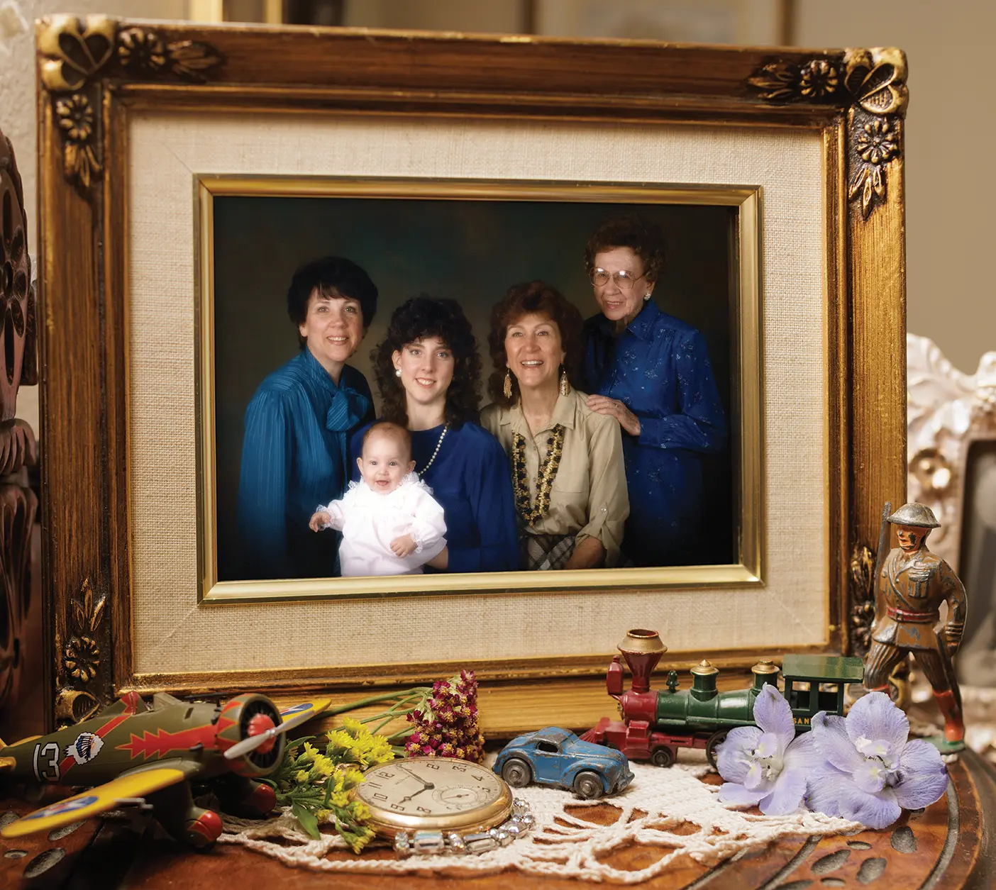 In an ornate, gilded picture frame set on a table, surrounded by various trinkets like a toy airplane and an old-fashioned stopwatch, four women and a baby, all part of the same family line, smile and pose in a photo.
