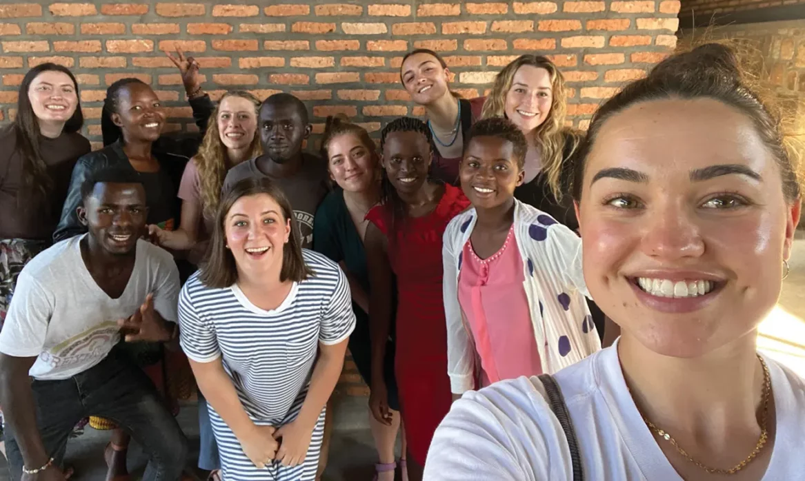 A group of female BYU students poses for a selfie with Rwandan peers in front of a brick building.