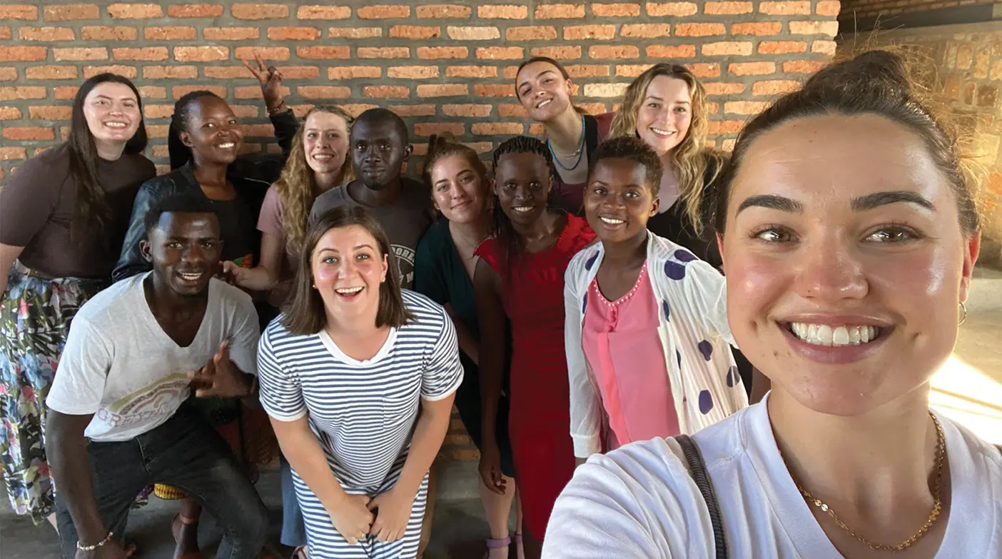 A group of female BYU students poses for a selfie with Rwandan peers in front of a brick building.