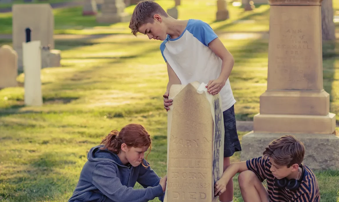 In a cemetery, three teens create an impression by rubbing chalk or charcoal on a paper pressed to a headstone.