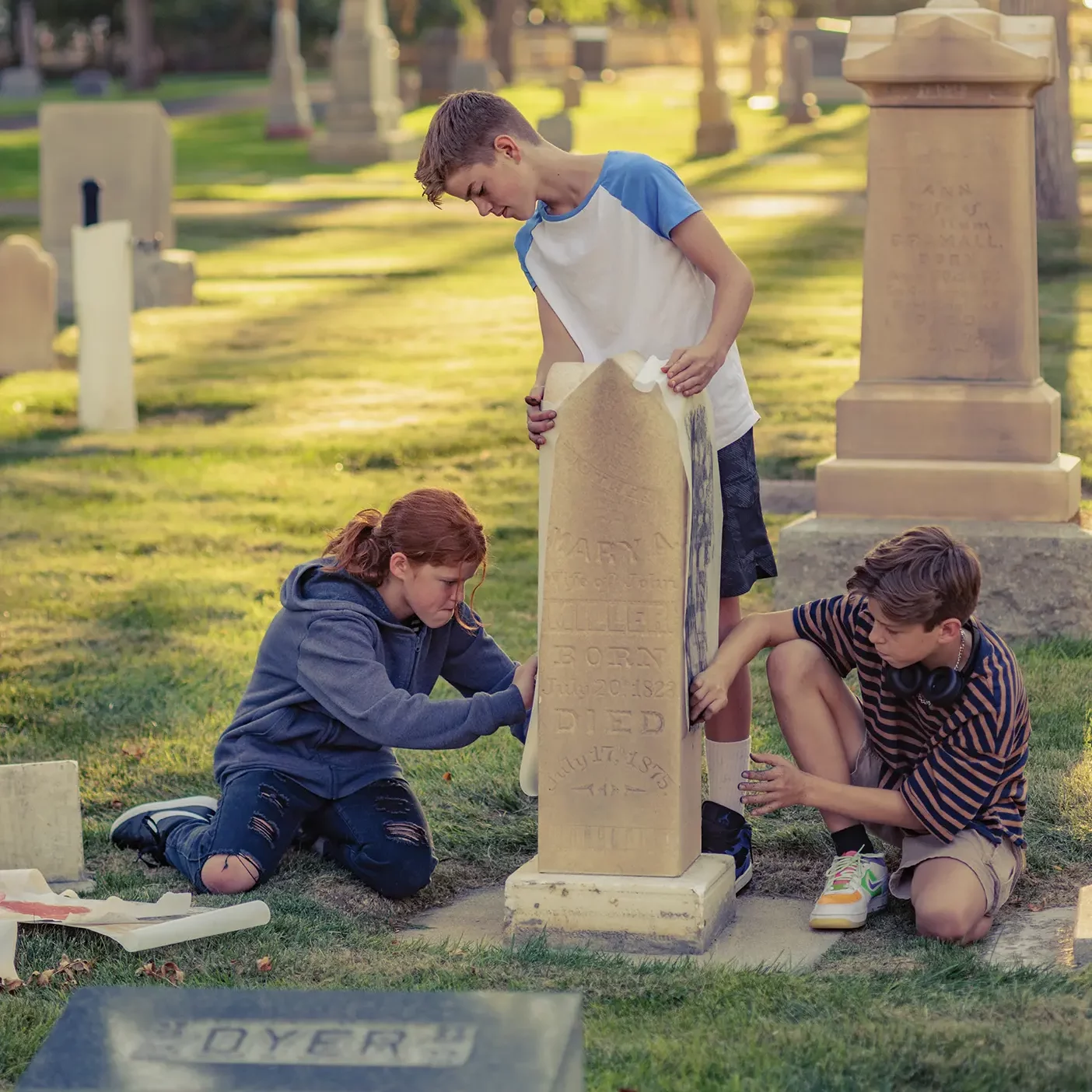 In a cemetery, three teens create an impression by rubbing chalk or charcoal on a paper pressed to a headstone.