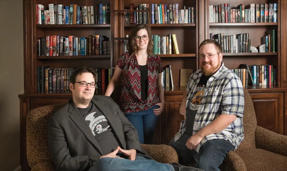 Brandon Sanderson poses with two of his former students, Charlie Holmberg and Brian McClellan, in front of a dark wooden bookcase.