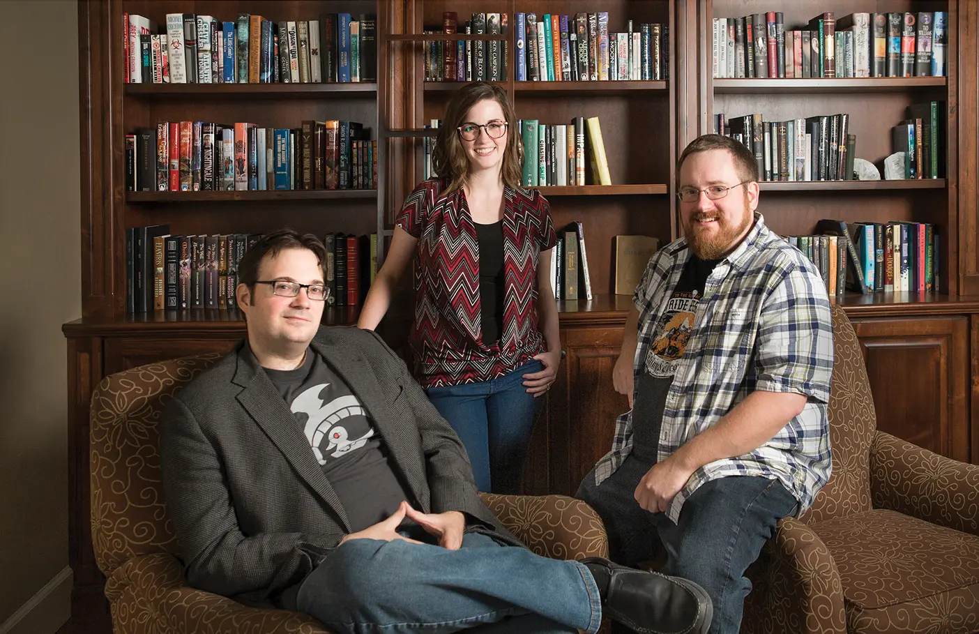 Brandon Sanderson poses with two of his former students, Charlie Holmberg and Brian McClellan, in front of a dark wooden bookcase.