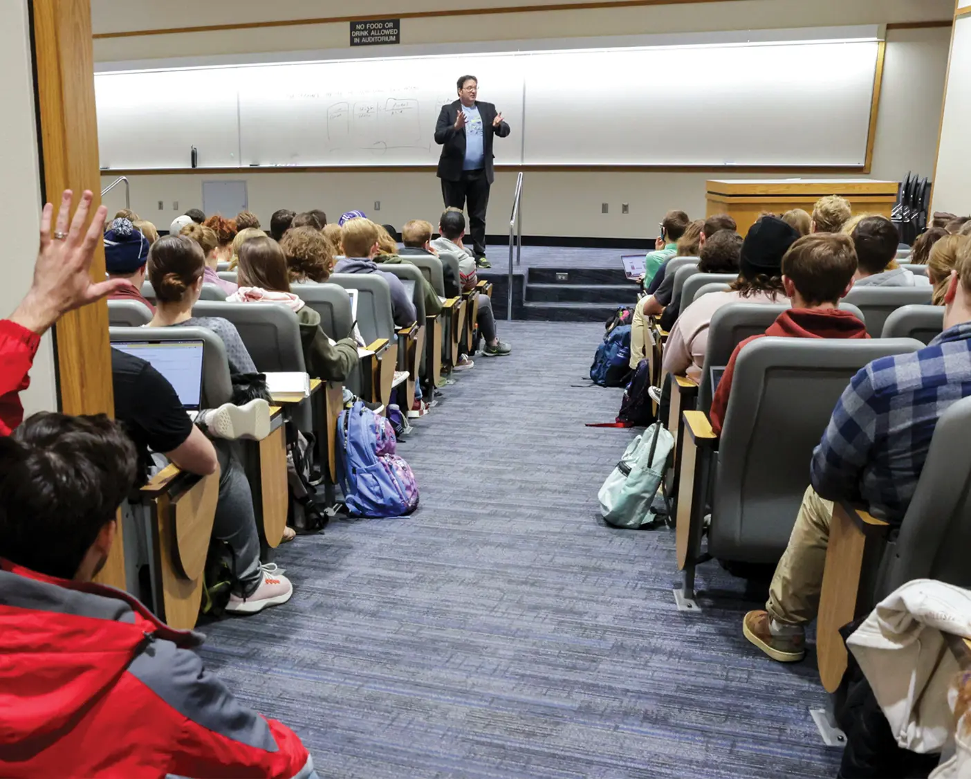 Brandon Sanderson stands at the front of a packed lecture hall. The hall is so full that some students sit in the entrance and hallway.