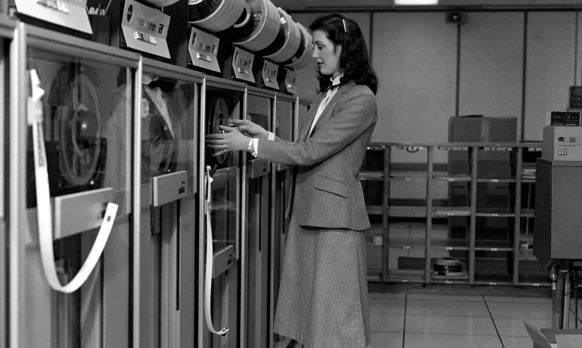 Woman dressed in long skirt and blazer adjusting early computer.