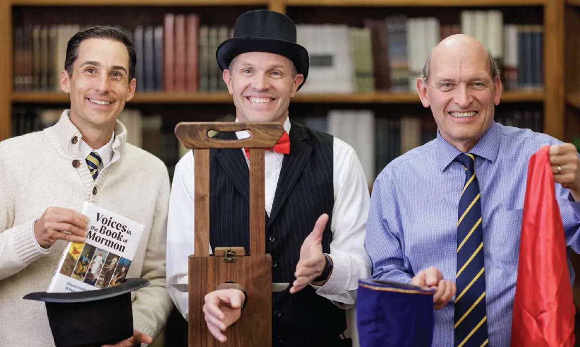 John Hilton III pulls his book "Voices in the Book of Mormon" from a hat, Devan Jensen demonstrates a guillotine magic trick, and Alex Baugh holds other magic props.
