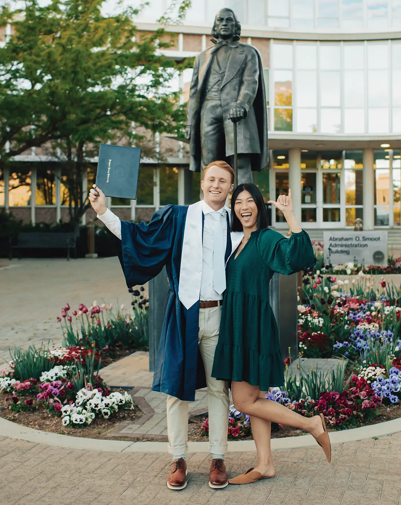 Samuel and Valerie Blackburn stand in front of a statue of Brigham Young, and Samuel wears graduation robes and holds a diploma.
