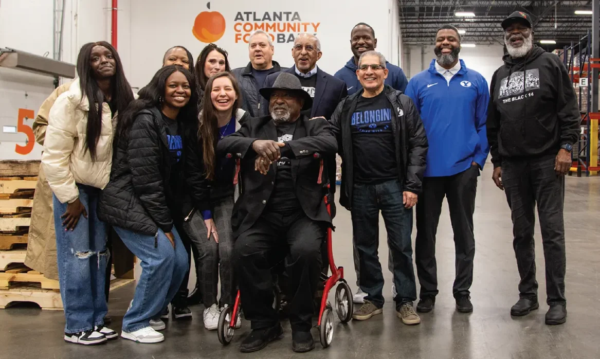 A group of volunteers, including students and a Black man in a wheel chair, pose for a photo in a food bank warehouse.
