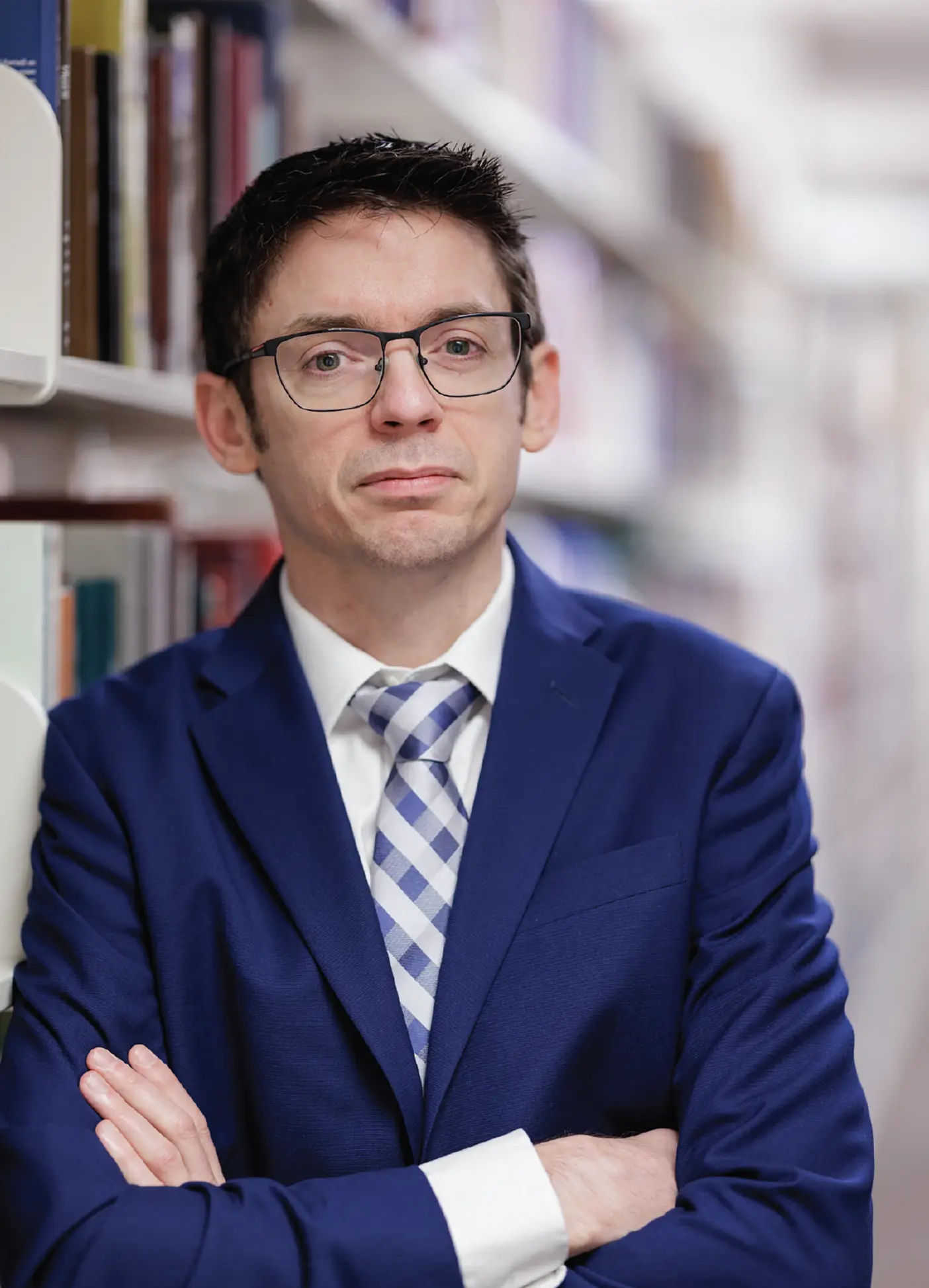 A man with glasses in front of a long book shelf looks at the camera.