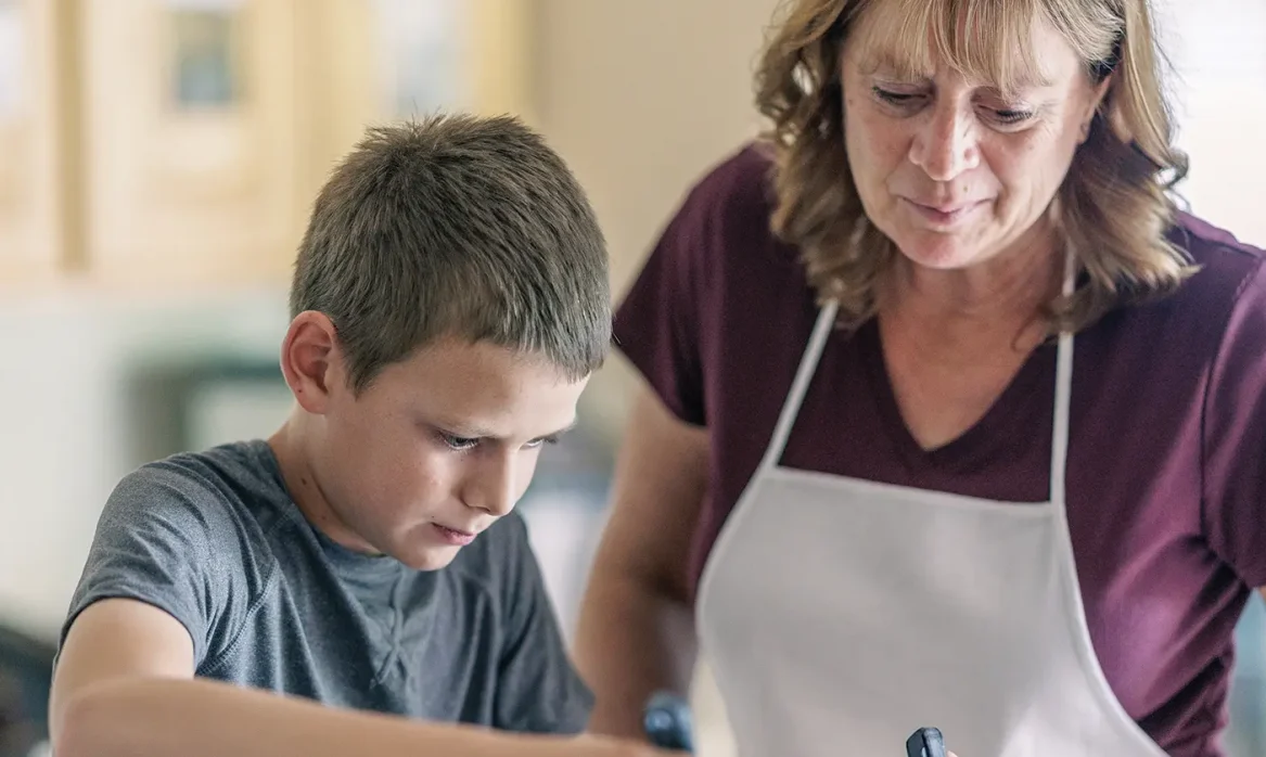 A young teenage boy uses to flip pancakes as his mother or grandmother observes.