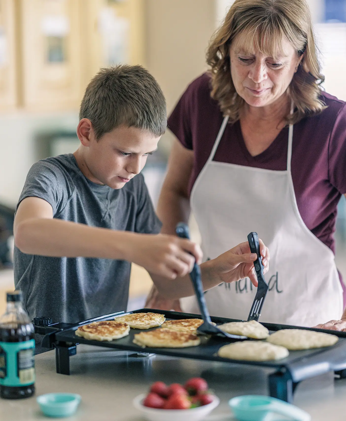 A young teenage boy uses two spatulas to flip pancakes as his relative offers motherly advice.