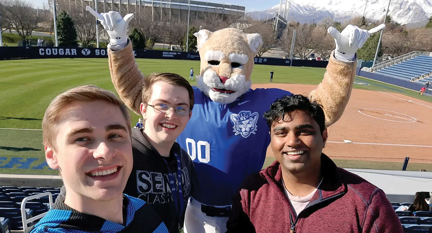 Three students with BYU mascot Cosmo at a BYU baseball game.