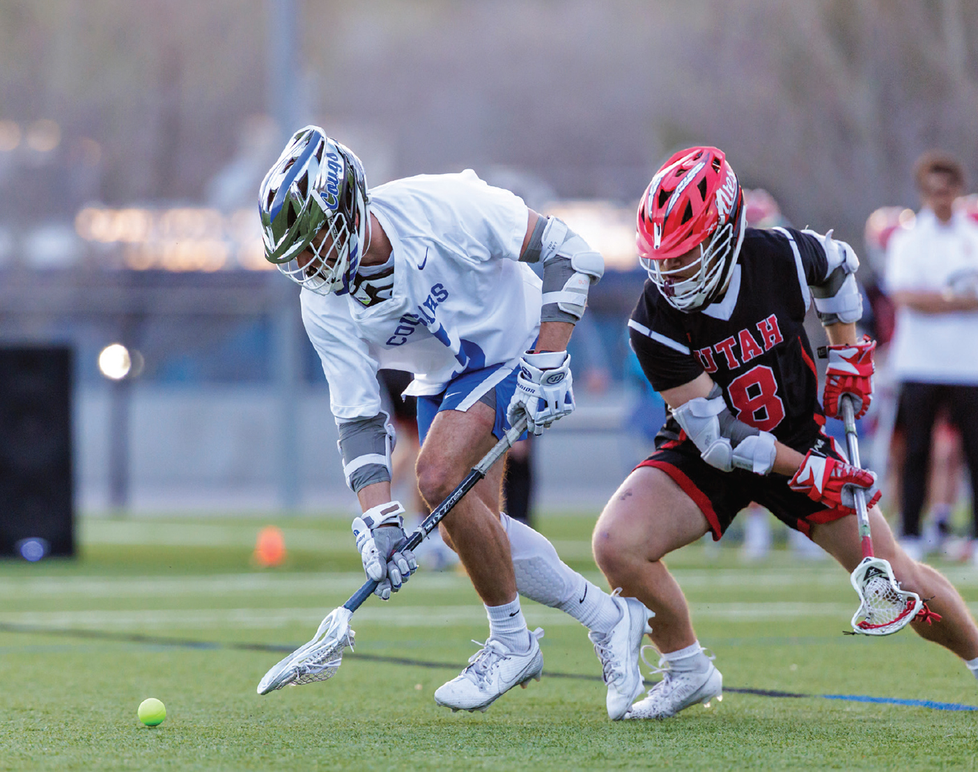 Two men's lacrosse players chase a ball across a field.