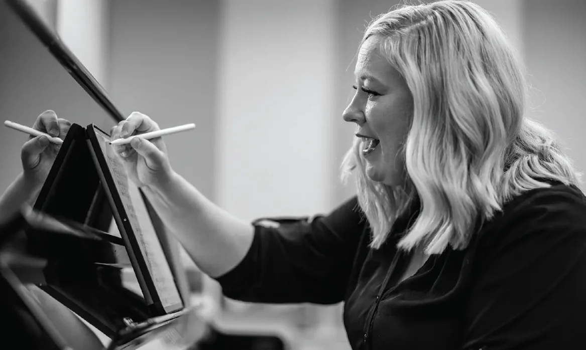 Black and white photo shows female professor at the piano, making notes with a stylus on a digital tablet displaying sheet music.