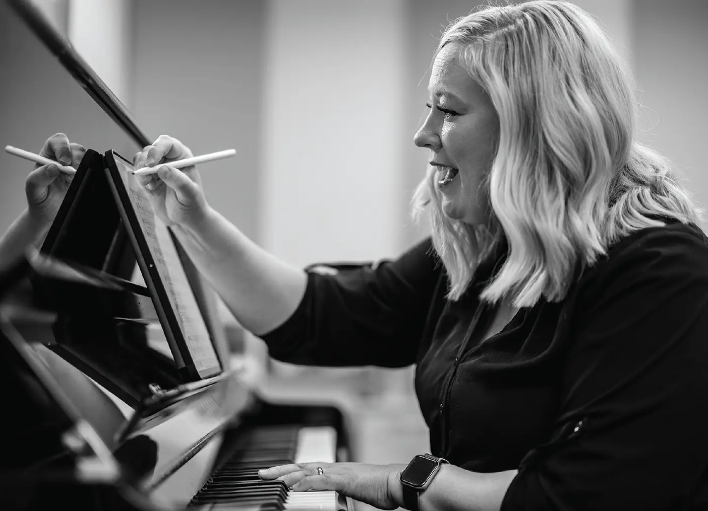 Black and white photo shows female professor at the piano, making notes with a stylus on a digital tablet displaying sheet music.