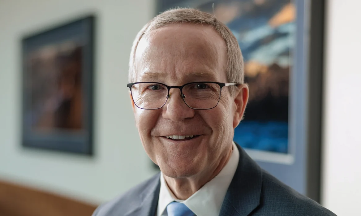 BYU school of medicine dean Mark Ott, an older man wearing a suit and glasses, stands in front of a painting in an office building.