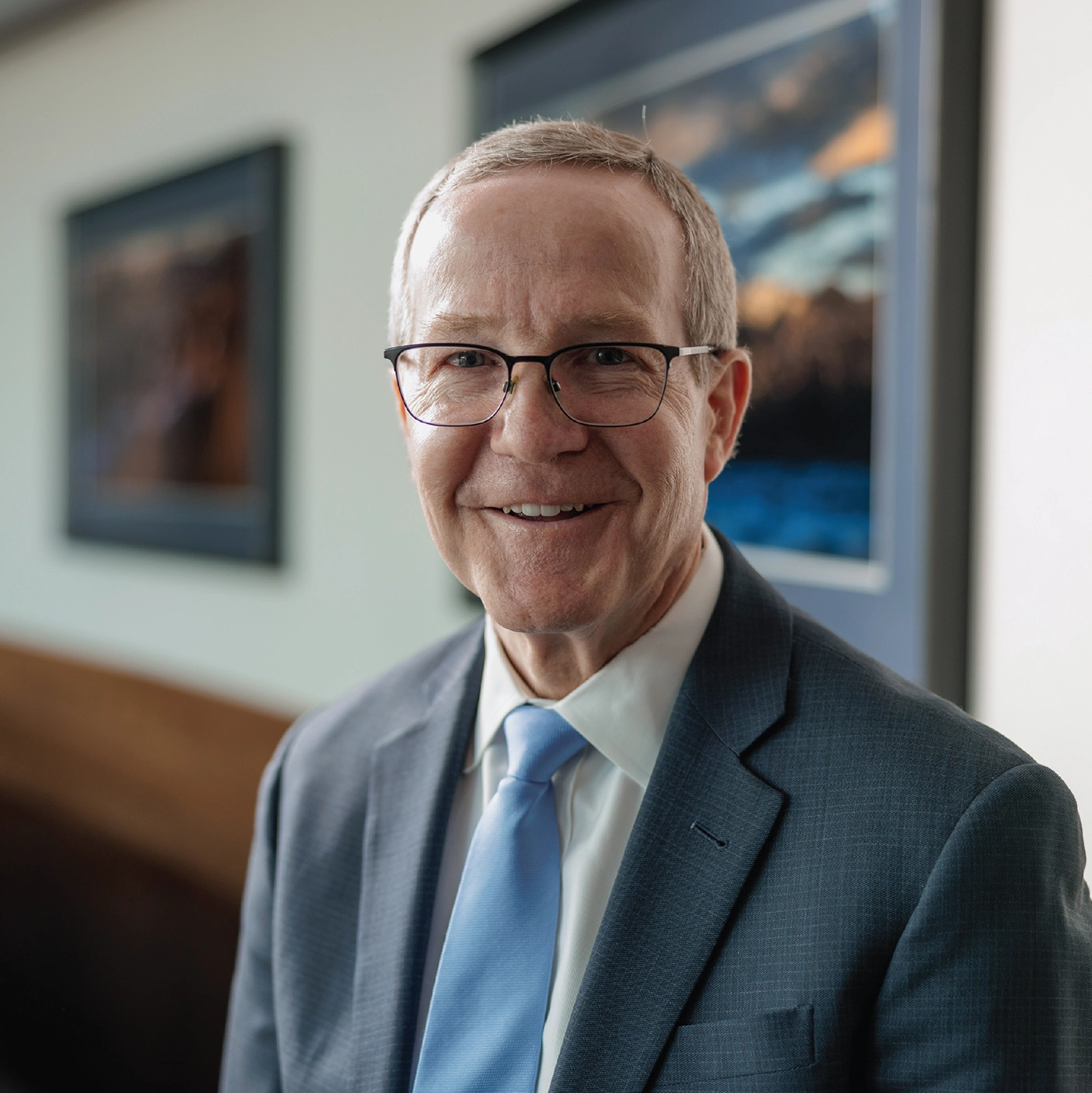 An older man in a suit wearing glasses smiles in front of a painting