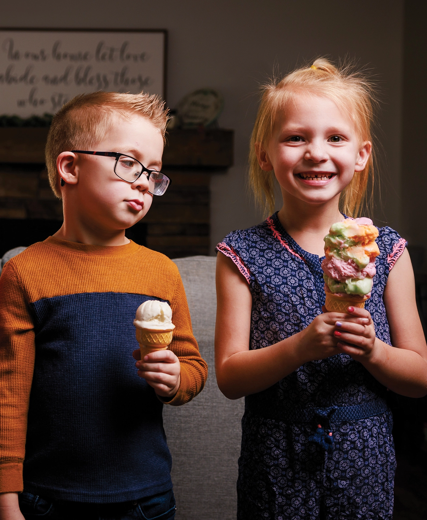 A young boy holding a one-scoop ice cream cone looks at his sister who has three scoops.