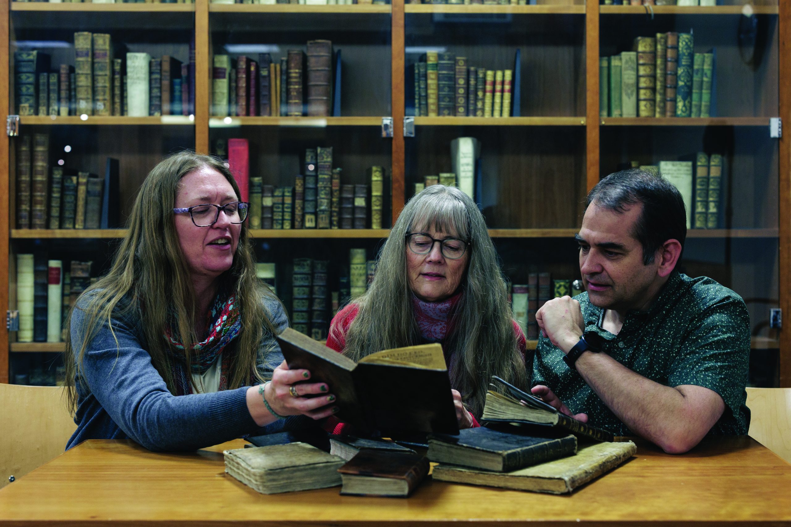 Two women and a man in a library sit at a table full of books and read together.