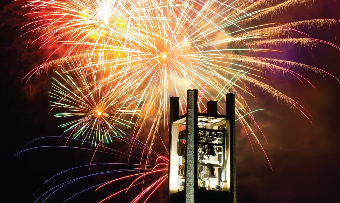 Multi-colored fireworks in warm tones explode behind the bell tower on BYU campus.