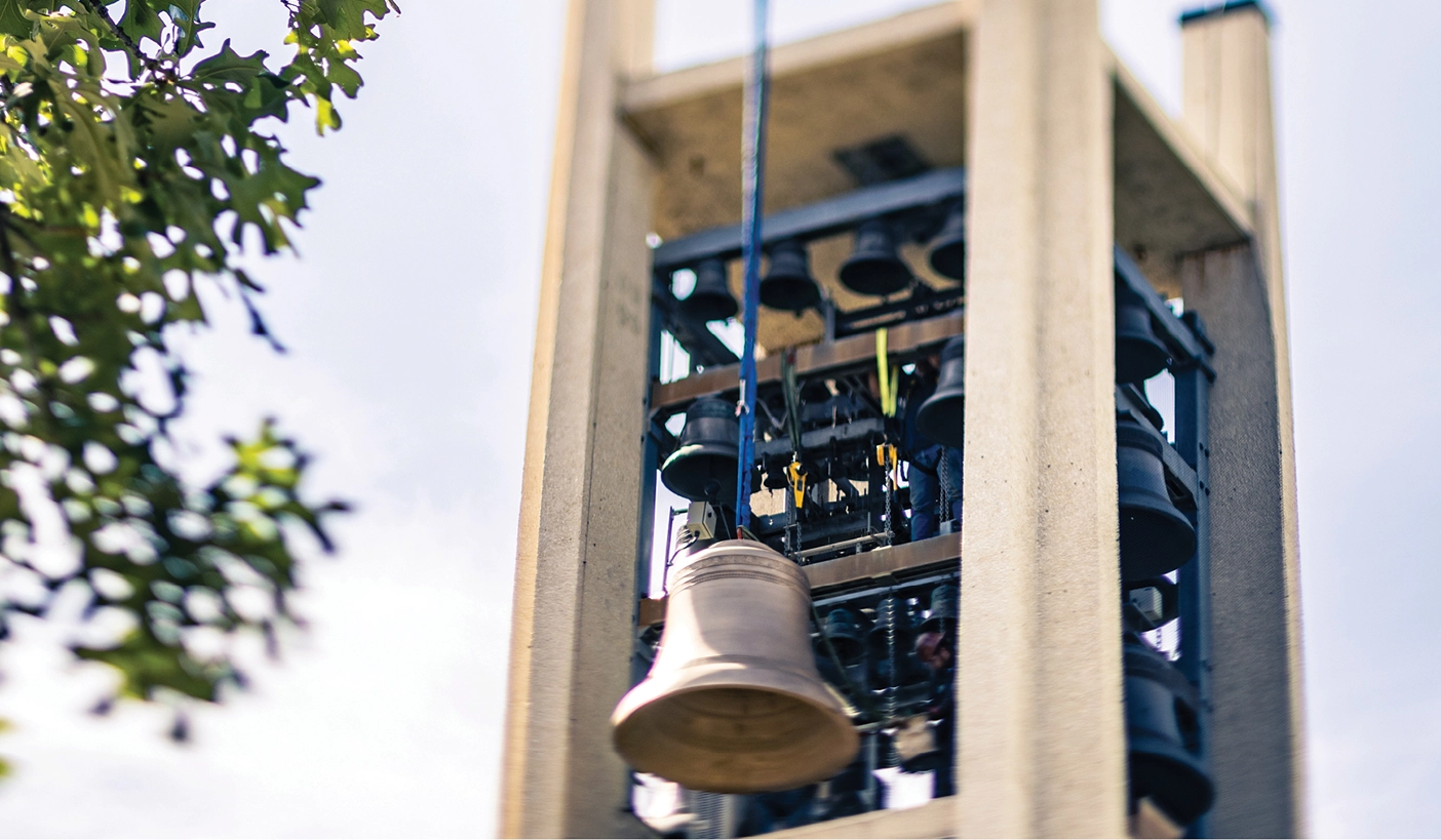 A large bell hangs from a crane in front of BYU's bell tower.