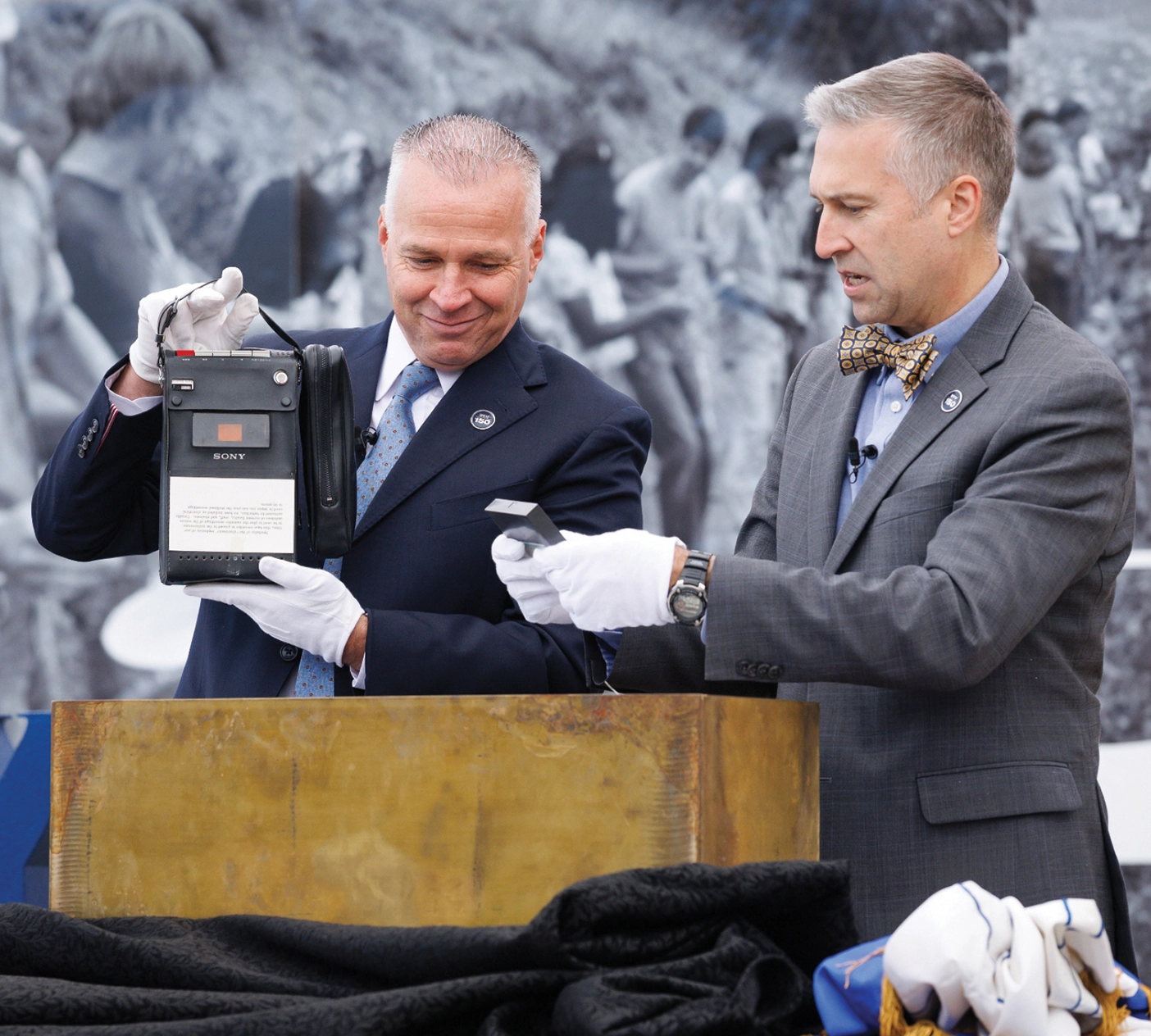 President C. Shane Reese and Richard Anderson stand next to a bronze box with white gloves, each holding historical items.