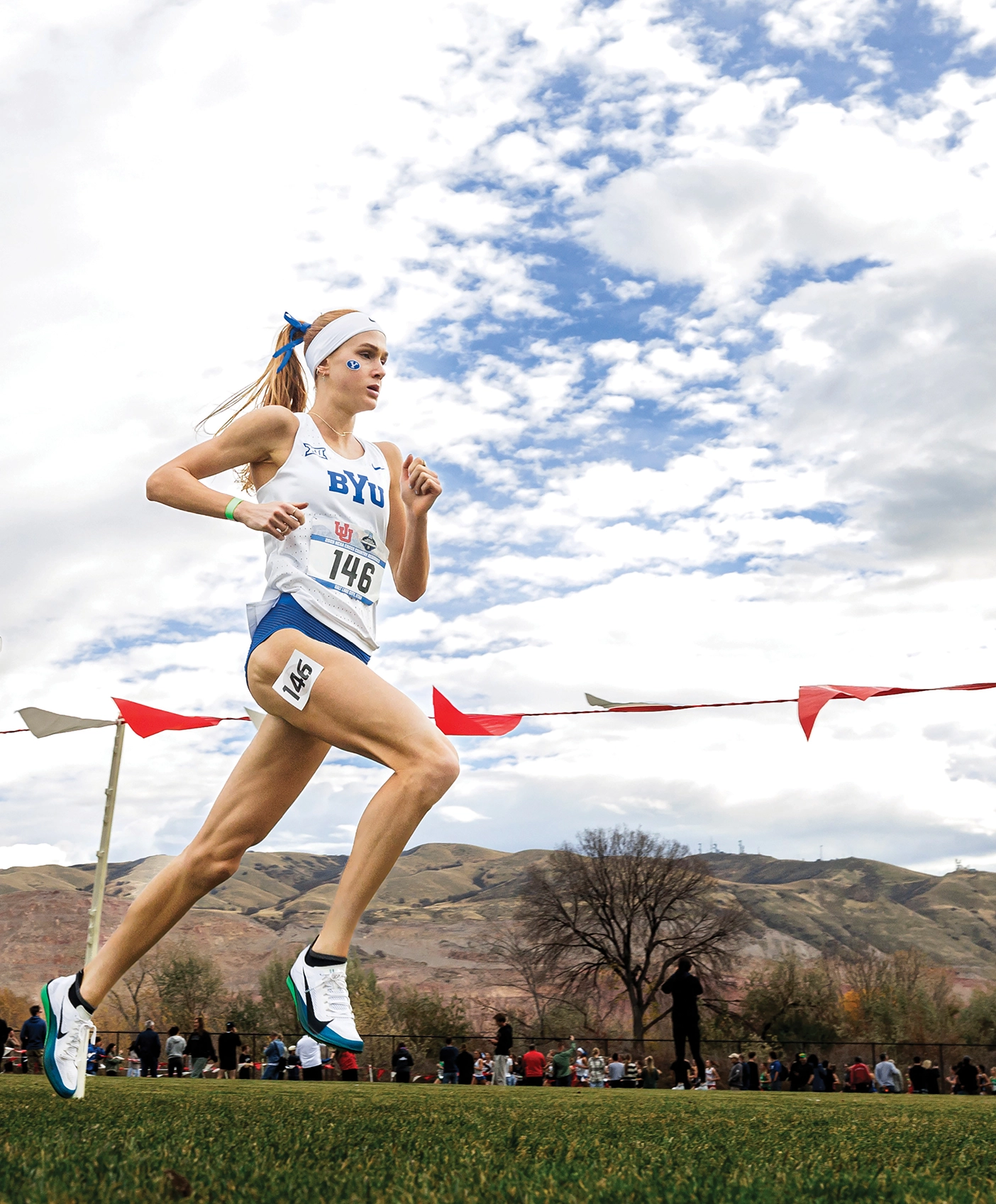 Jane Hedengren, a cross country and track athlete, runs in a race on the grass against a blue cloudy sky.