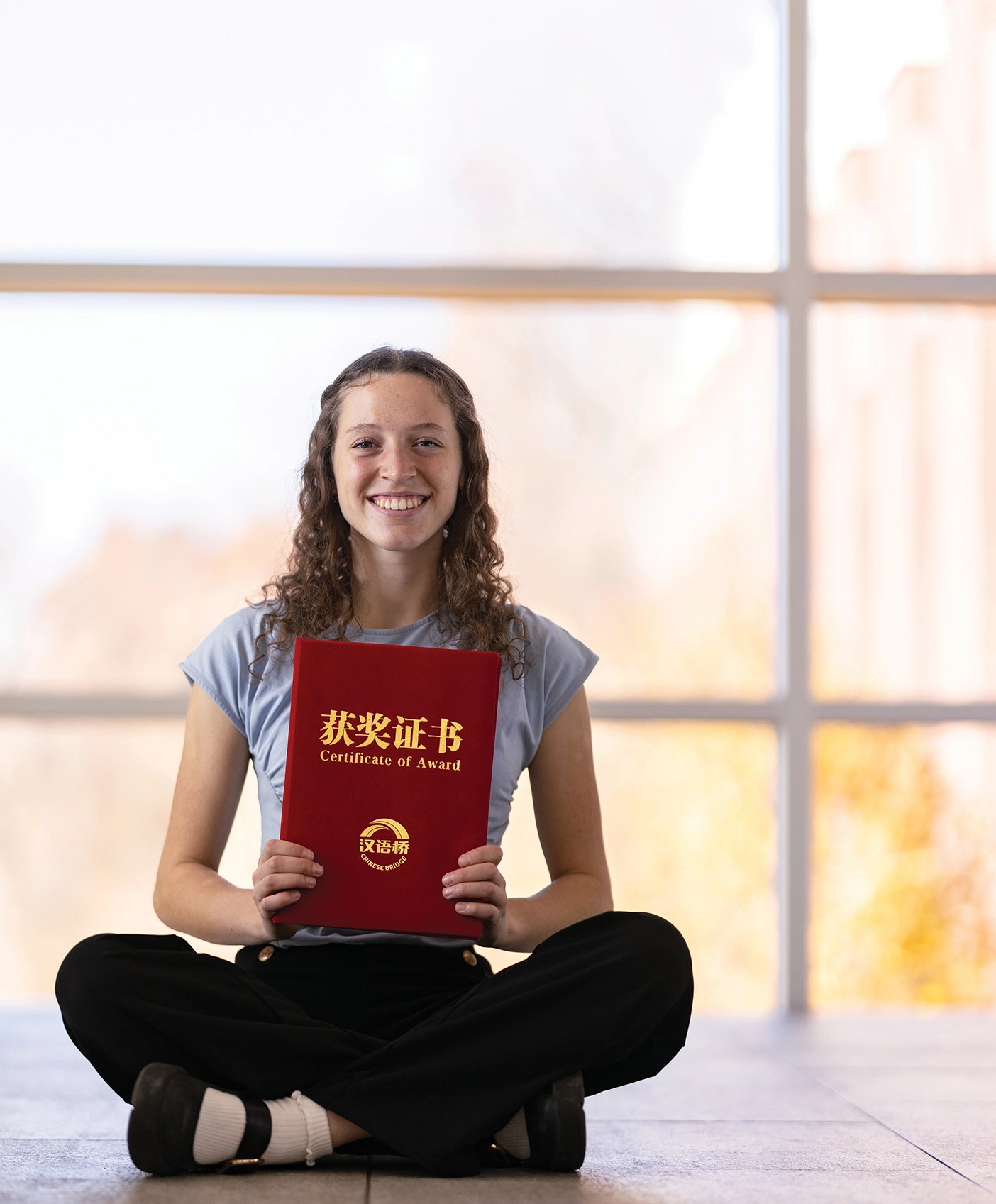 Ashley Breinholt sits in front of a window with a red Certificate of Award printed with gold Chinese characters in her hands.