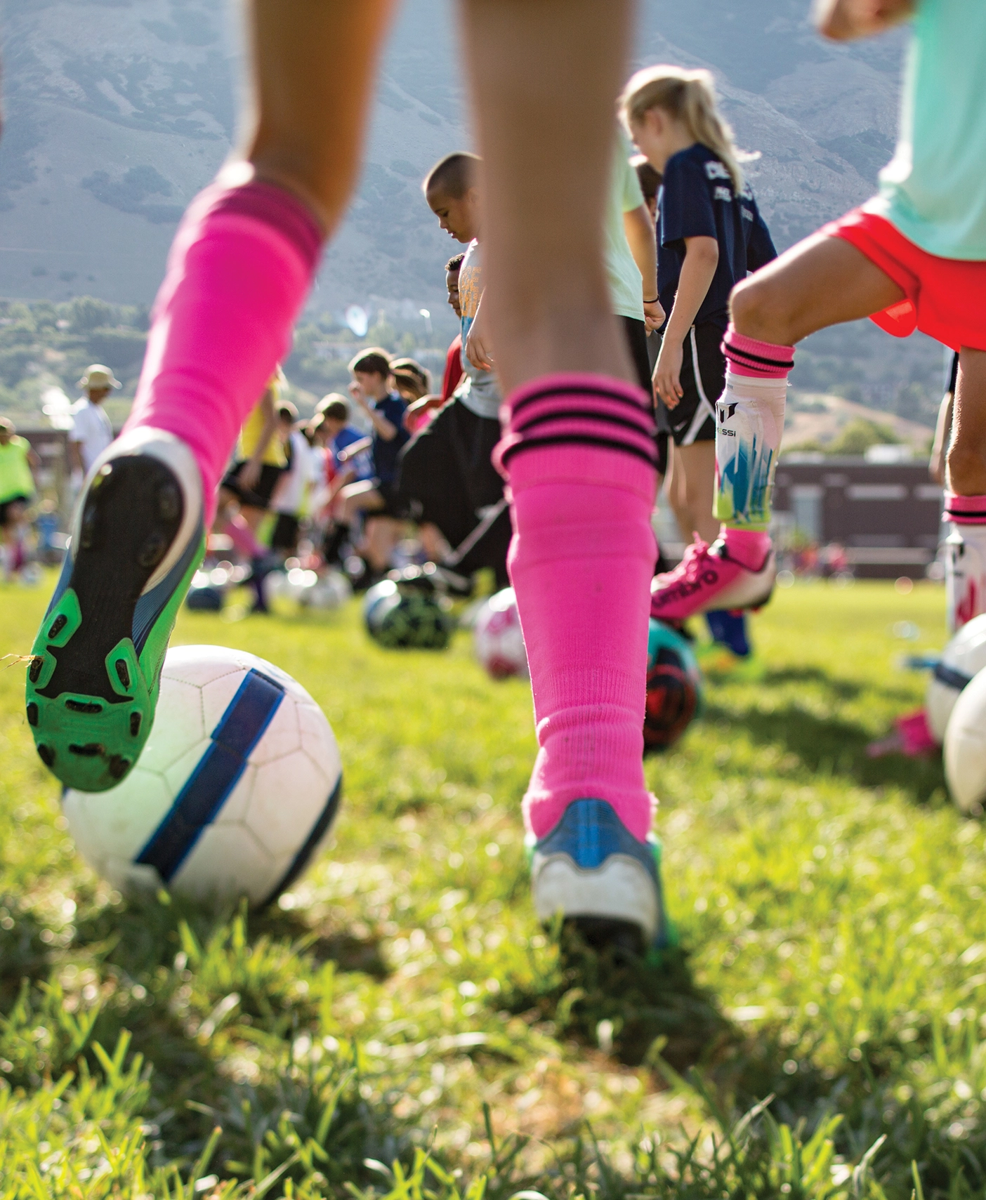 Low-angle photograph of children practicing soccer on a grassy field. In the foreground, a child wearing bright pink knee-high socks and cleats rests one foot on a soccer ball. Other children stand nearby with their own balls, partially out of focus, suggesting a group drill or warm-up. Mountains and buildings are visible in the background, indicating an outdoor field on a sunny day.