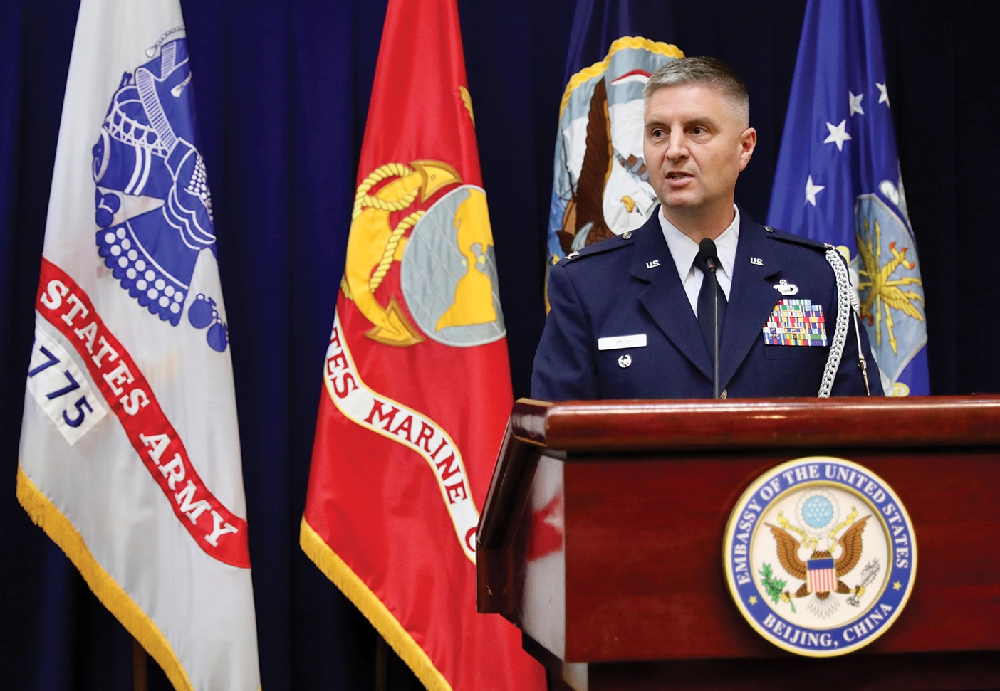 Steven Smith speaks at the US Embassy in Beijing, standing at a podium in front of flags from various countries.