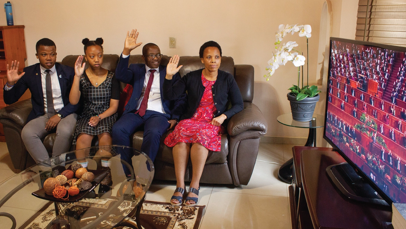 A family in Africa sits on a couch together, raising their arms in unison to sustain church leaders as they watch general conference on the television.