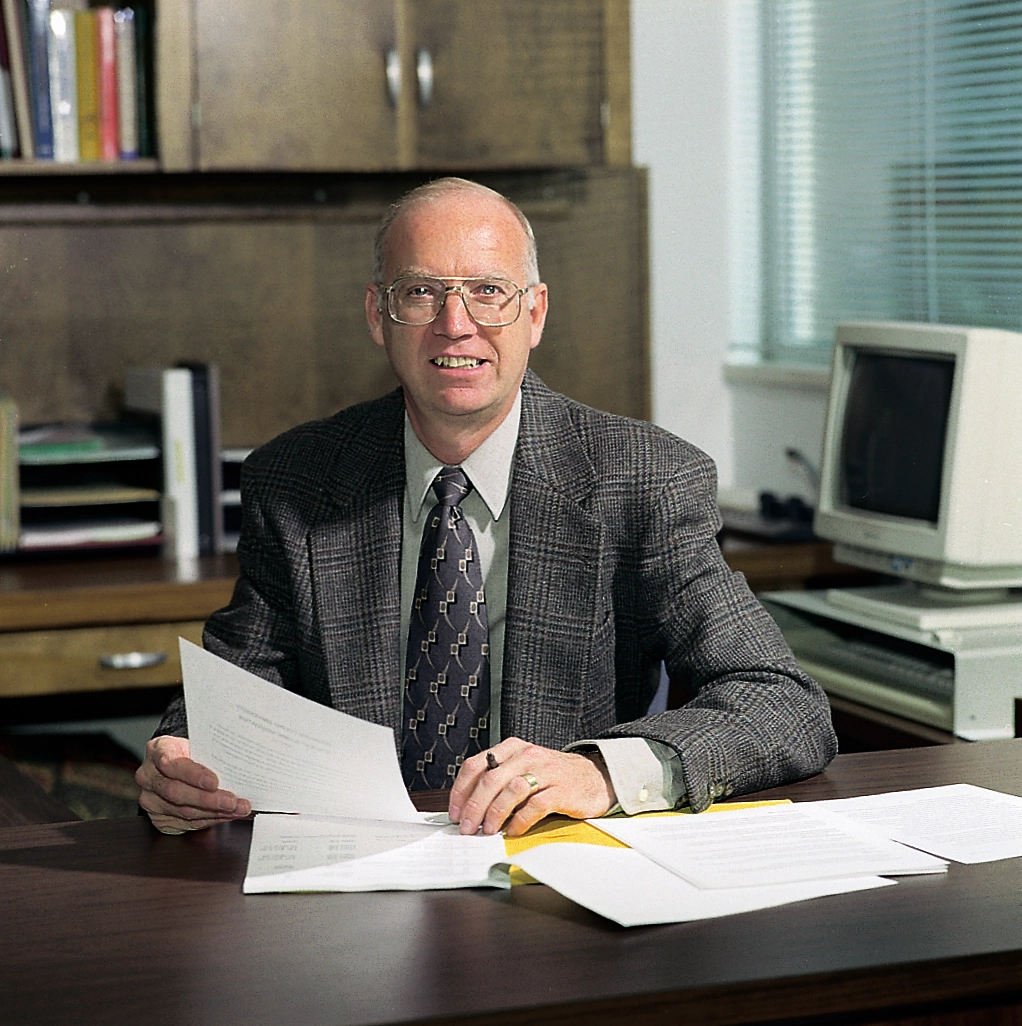A man in a suit sits at an office desk holding a document, with bookshelves and a computer behind him.