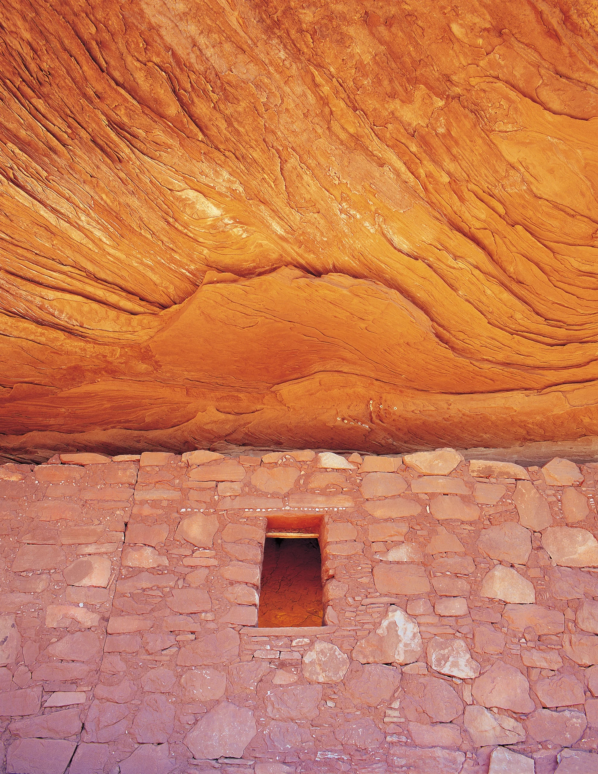 Stone wall with a small square opening beneath a broad, rippling orange sandstone ceiling.