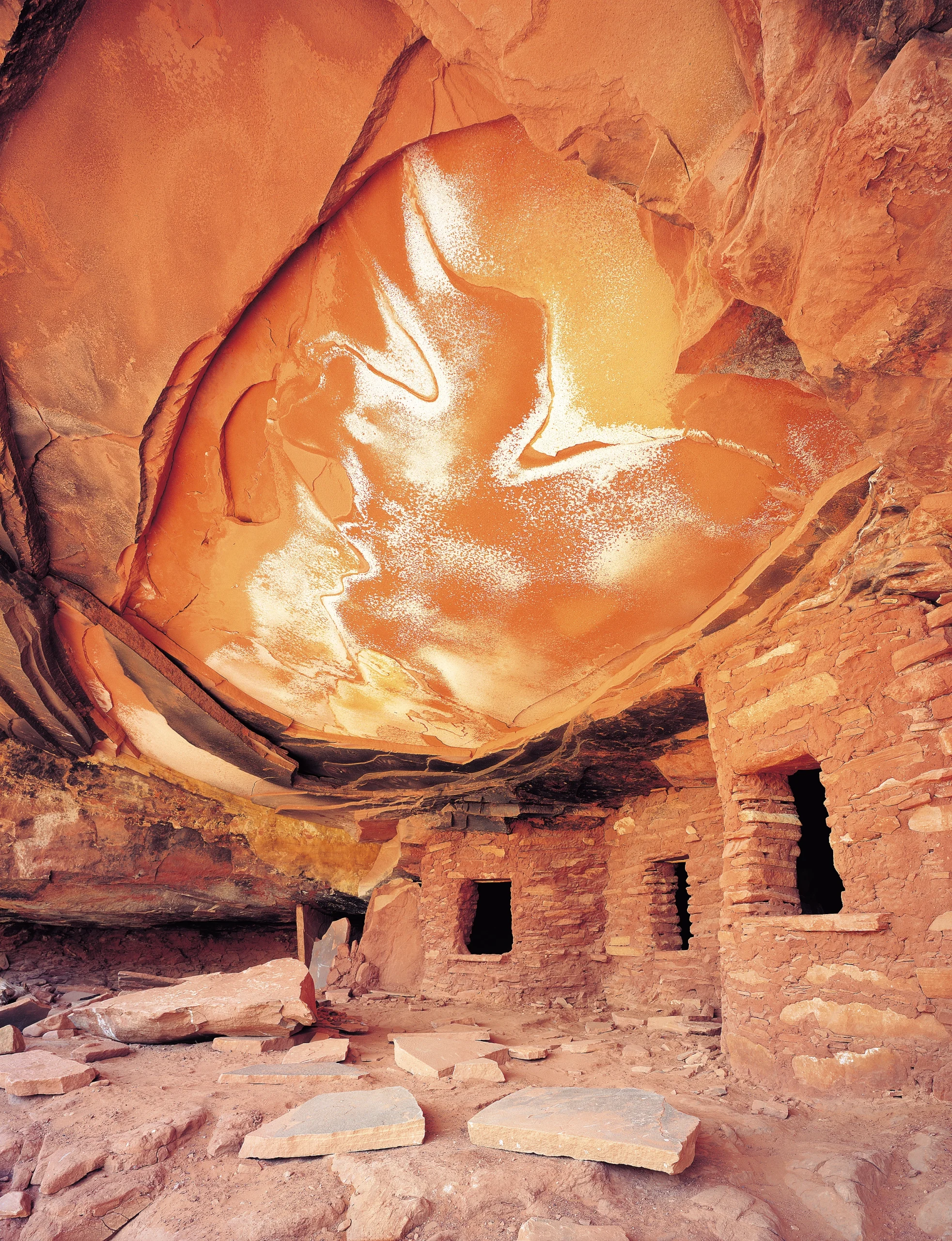 Small stone dwellings beneath a dramatic curved sandstone alcove illuminated in glowing orange and cream tones.