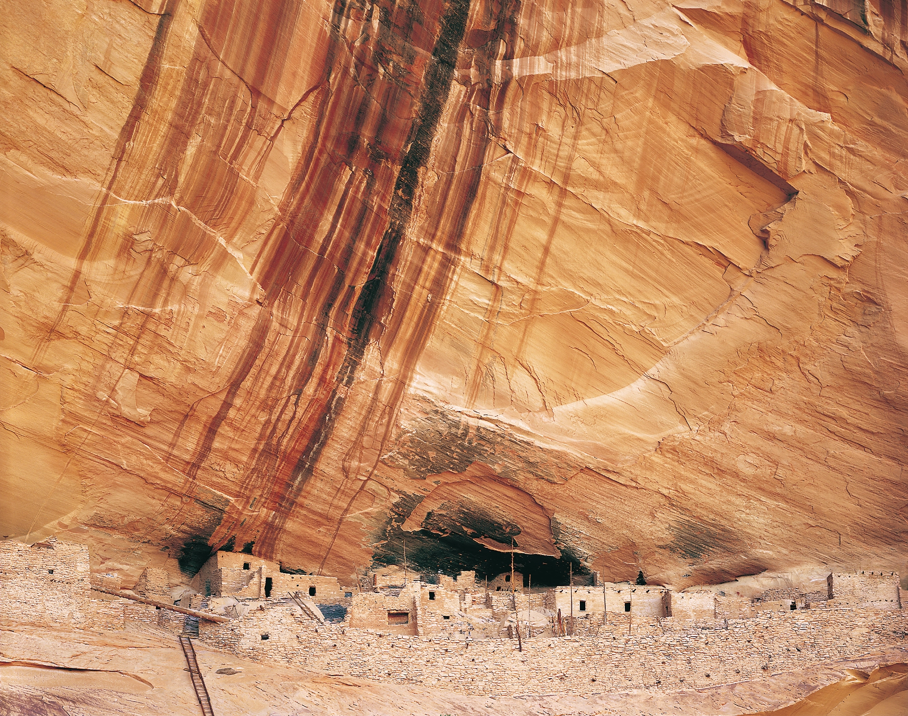 A long line of pale stone dwellings sheltered at the base of a towering, vertically streaked sandstone wall.