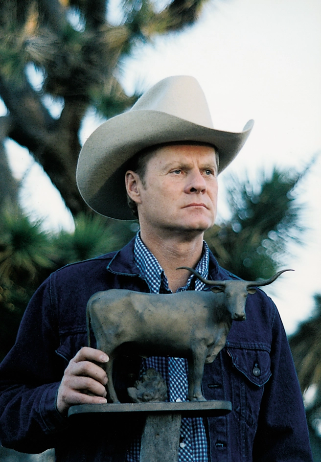 A rancher in a white cowboy hat holds a small bronze sculpture of a longhorn steer.