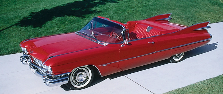 Bright red vintage convertible with prominent tail fins parked on a paved path beside grass.