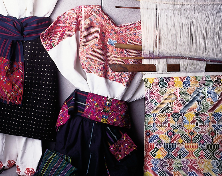 Museum display of vividly patterned woven garments, fabric panels, and a loom.