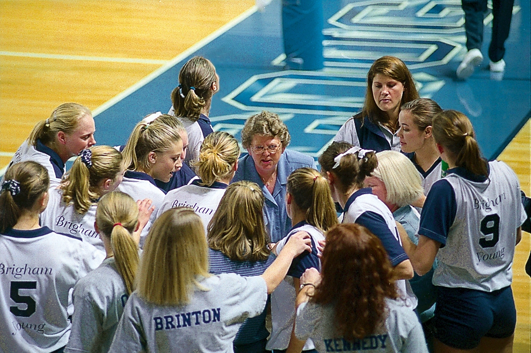 Women’s volleyball players form a tight huddle around their coach on an indoor court.