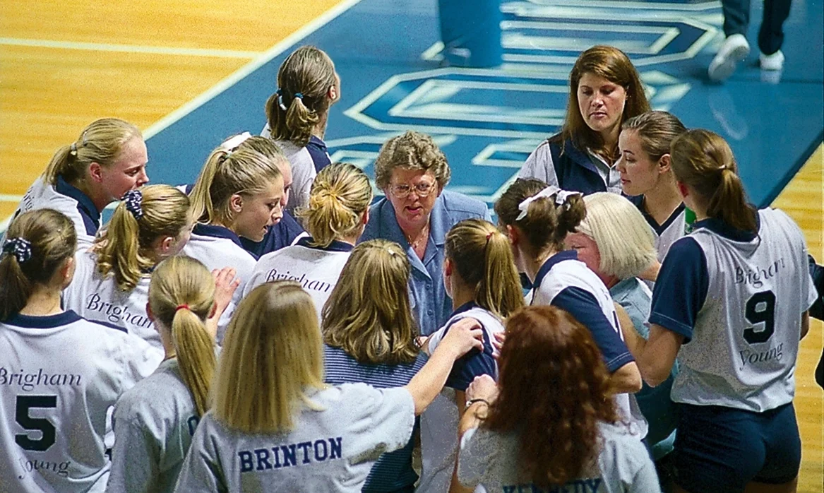 Women’s volleyball players form a tight huddle around their coach on an indoor court.