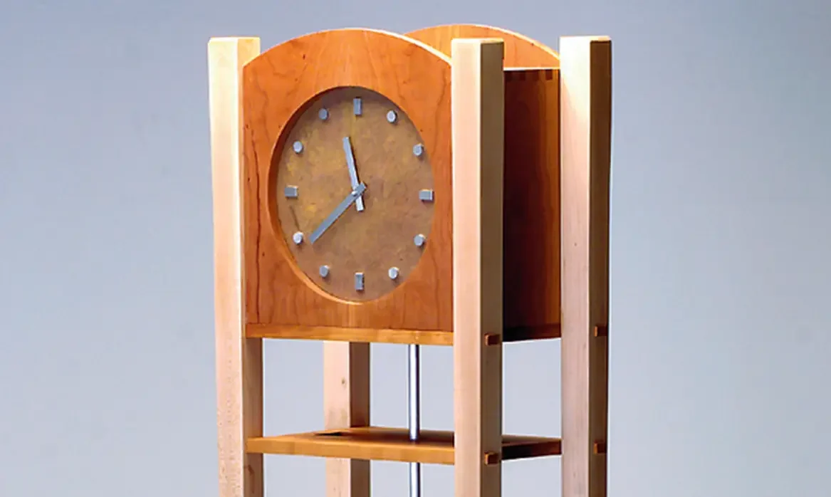 Tall wooden grandfather-style clock designed by Drew N. Soderborg, with four slender legs, a rectangular frame, a round clock face near the top, and a long pendulum hanging below.