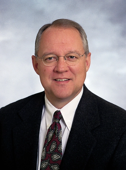 Formal color headshot of an older man wearing glasses, a dark suit, and a patterned tie.