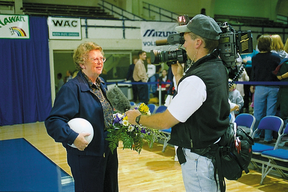 A woman holding a volleyball speaks with a camera operator beside an indoor volleyball court.