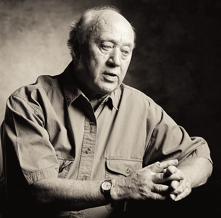 Black-and-white portrait of an older man seated with his hands clasped, wearing a casual button-front shirt.
