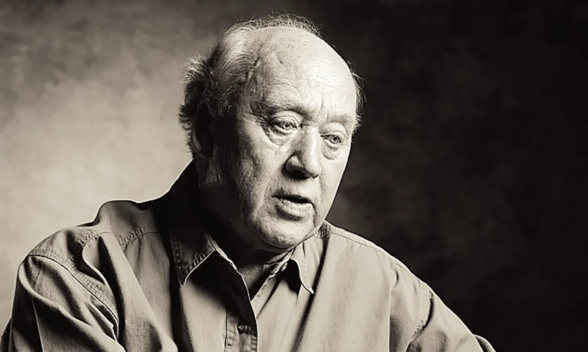 Black-and-white portrait of an older man seated with his hands clasped, wearing a casual button-front shirt.